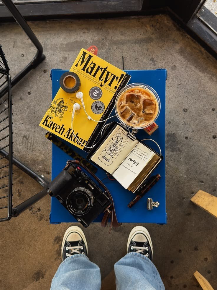 Blue table with a yellow book, iced coffee, notebook and camera, seen from above