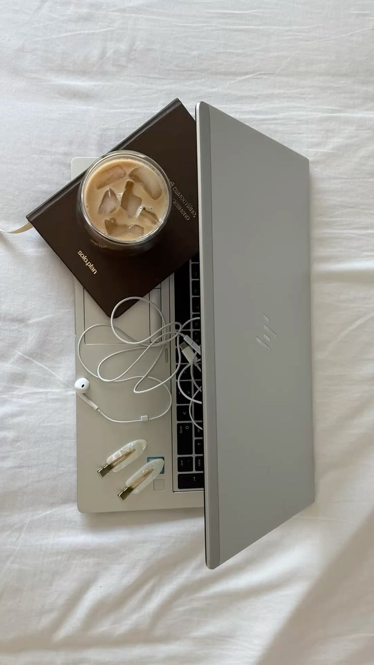 Laptop, iced coffee, planner and earphones on a white bed